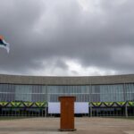 Fachada da Câmara Legislativa do DF com bandeiras e pódio vazio, representando cerimônia cívica questionada em crise econômica.