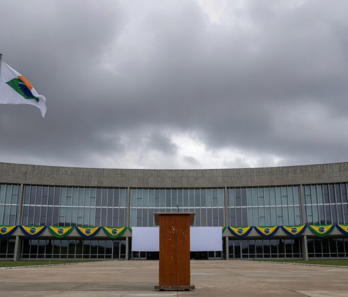 Fachada da Câmara Legislativa do DF com bandeiras e pódio vazio, representando cerimônia cívica questionada em crise econômica.