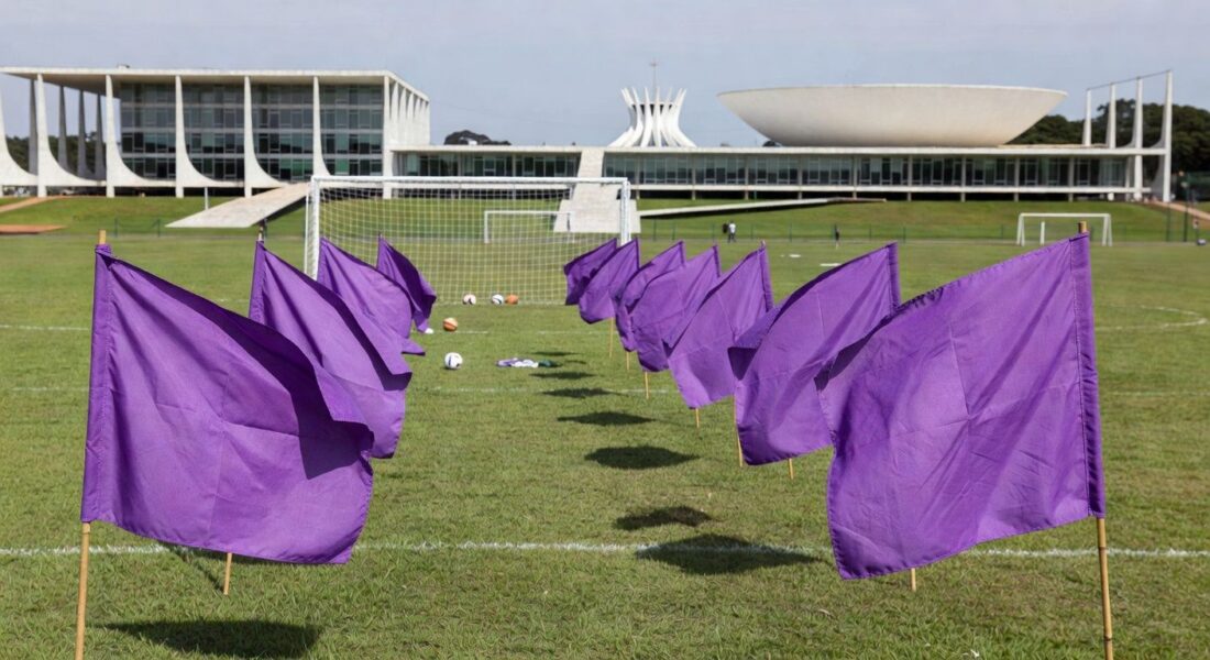 Auditório em Brasília com banners roxos contra feminicídio, ao fundo Congresso Nacional.