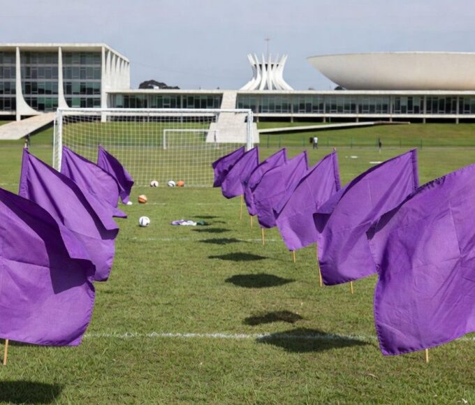 Auditório em Brasília com banners roxos contra feminicídio, ao fundo Congresso Nacional.