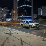 Cena de rua em Águas Claras com viatura policial e fita de isolamento após incidente de assalto.
